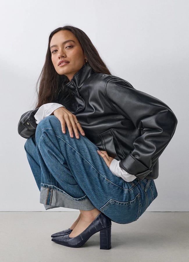 A woman with long dark hair squats, wearing a black leather jacket, blue turned-up jeans, and sexy heeled shoes against a plain light background.