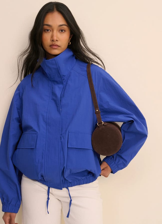 A woman in a bright blue jacket—a nod to the International Klein Blue colour trend—and white trousers stands against a light background, wearing a small, round brown shoulder bag and looking at the camera.