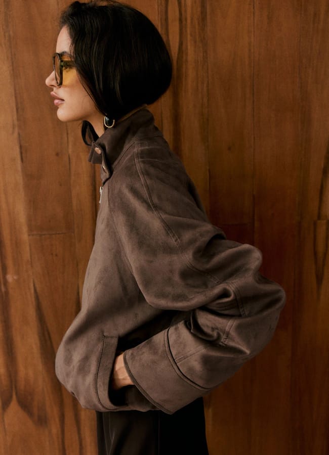 A person with short dark hair wearing glasses and a loose, lightweight brown jacket stands in profile against a wooden wall, with one hand in their pocket.