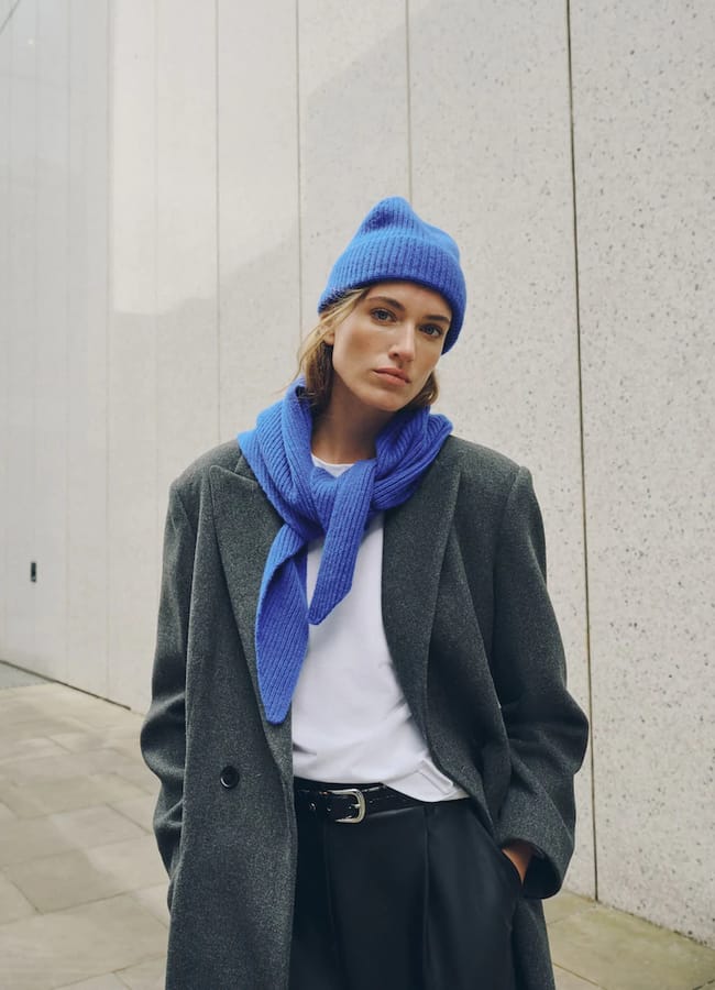 Person standing outside wearing an International Klein Blue beanie and jumper draped over shoulders, grey coat, white shirt, and black trousers, with hands in pockets against a light stone wall background. The striking colour of the beanie brings a bold accent to the look that stands out everywhere.