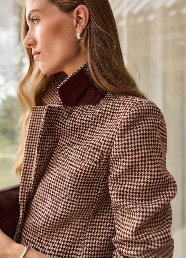 Woman with long wavy hair wearing a brown houndstooth-patterned coat with a dark collar, standing indoors and looking to the side near a window—capturing the essence of expensive fashion and classic style essentials.