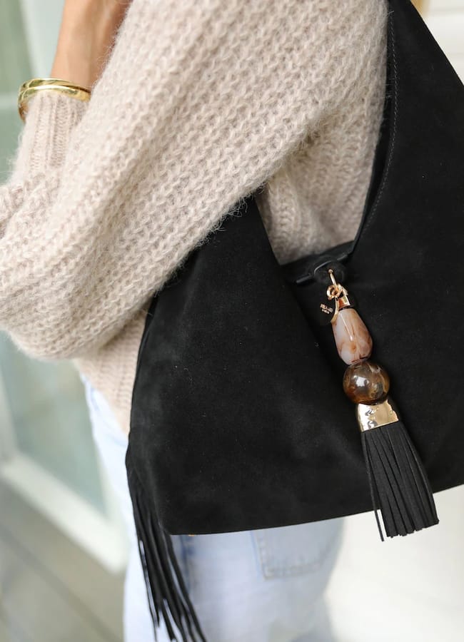 Person wearing a beige knitted jumper and light jeans holds a black suede handbag with a tassel and beaded charm—a perfect accessory for cool girls embracing autumn style. Only the torso and arm are visible.