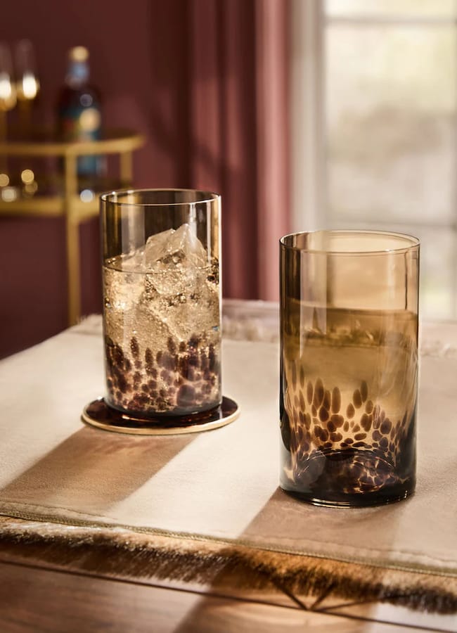 Two brown-tinted glasses with a dotted pattern sit on a table; one glass contains ice and a drink, the other is empty. Sunlight streams through a window, casting shadows and warm tones across the scene—an inviting display of chic homewares perfect for any modern space.