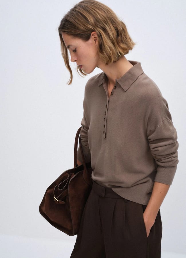A woman with short, wavy hair wears a taupe long-sleeved polo and brown trousers, holding a large brown bag—her look exuding the effortless style often seen in minimalist high street brands—as she stands against a plain light background, looking downward.
