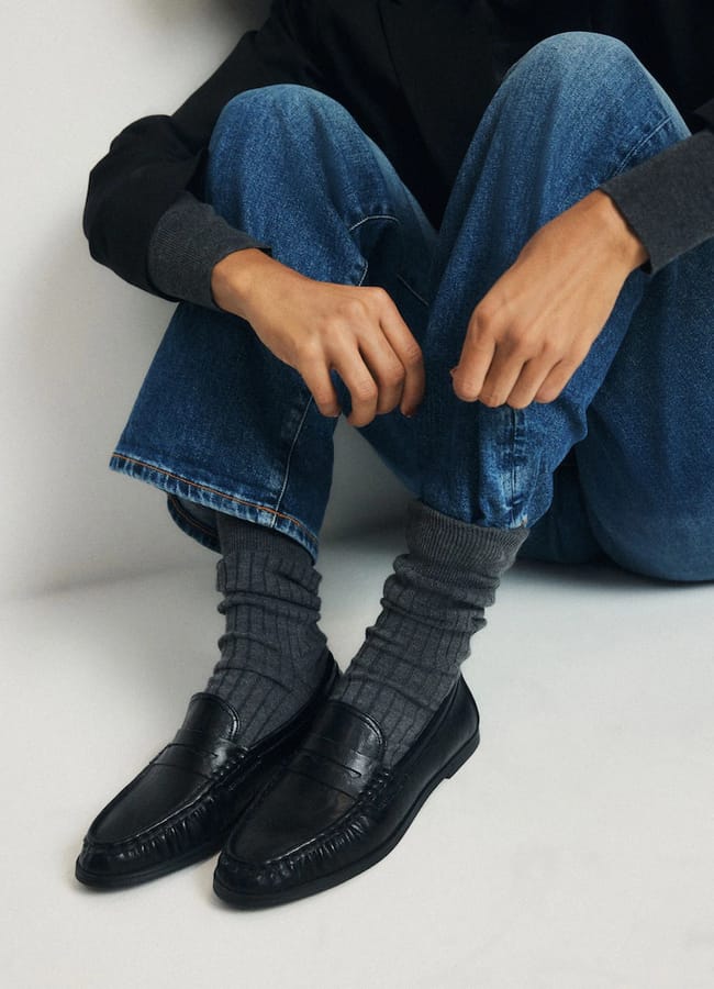 Person sitting on the floor with knees up, wearing blue jeans, a dark jacket, grey ribbed socks, and black loafers—showcasing some of the best women’s loafers; hands resting on legs.