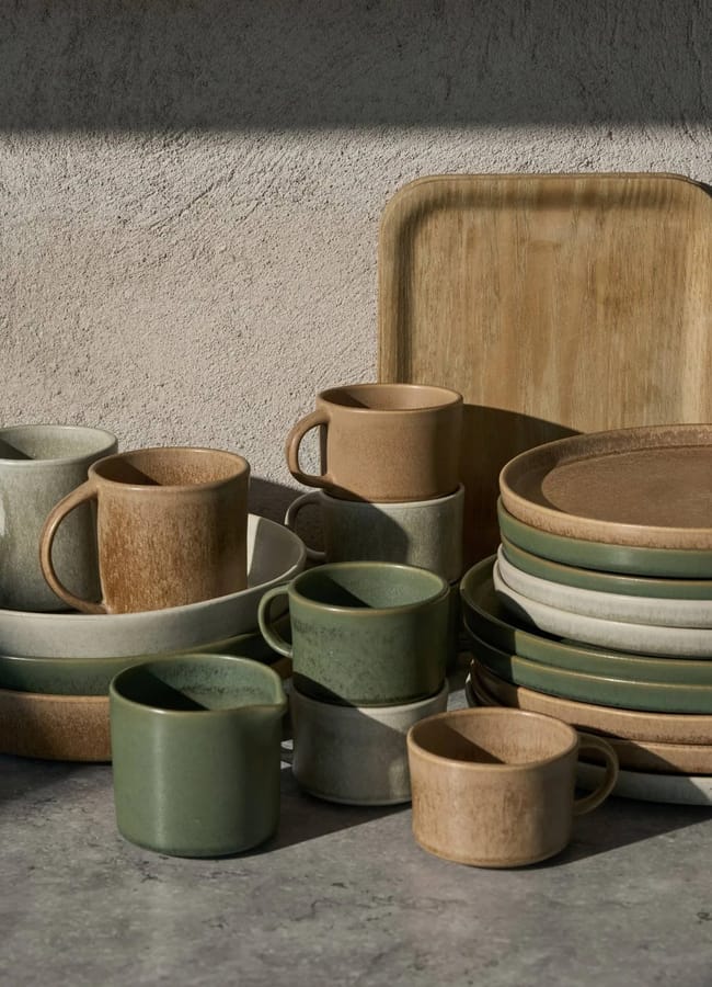 Assorted ceramic dishes, cups, and plates in earthy tones of brown, green, and beige are neatly stacked on a grey surface against a textured wall, lit by soft natural light—perfect chic homeware under £50 to elevate any table setting.