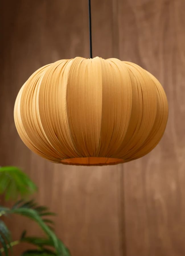 A round, pleated, golden-yellow fabric pendant lamp hangs from the ceiling, set against a wooden background with a green leafy plant in the lower left corner—a warm accent piece that beautifully complements caramel homeware.