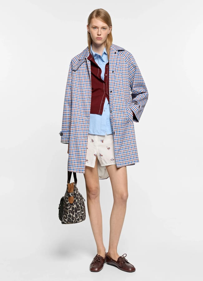 A person stands in a blue and red chequered lightweight jacket over a blue shirt and burgundy jumper, wearing white shorts with small prints, brown shoes, and holding a leopard print handbag. The background is plain white.