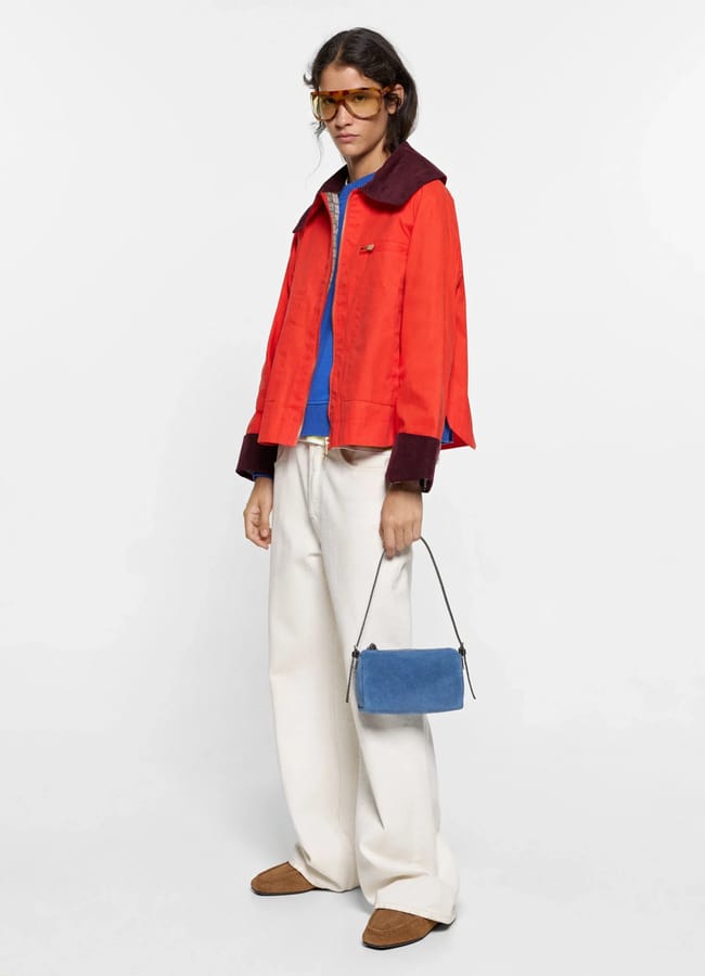 Person wearing a lightweight red jacket, blue shirt, white wide-leg trousers, brown shoes, brown sunglasses, and holding a small blue handbag, standing against a plain white background.