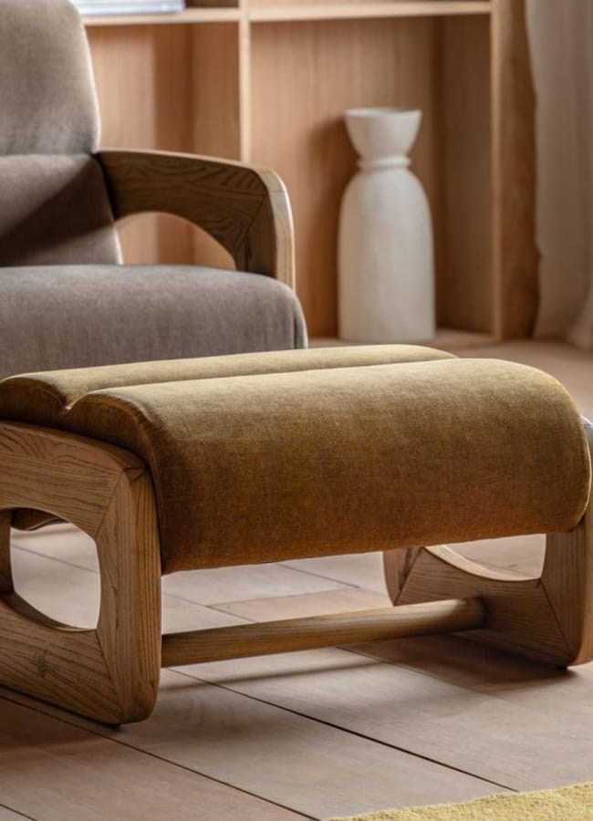 A modern ottoman with a curved wooden frame and mustard-yellow cushion sits on a wooden floor near a beige armchair and shelving unit adorned with caramel homeware, all in a cosy, well-lit room.