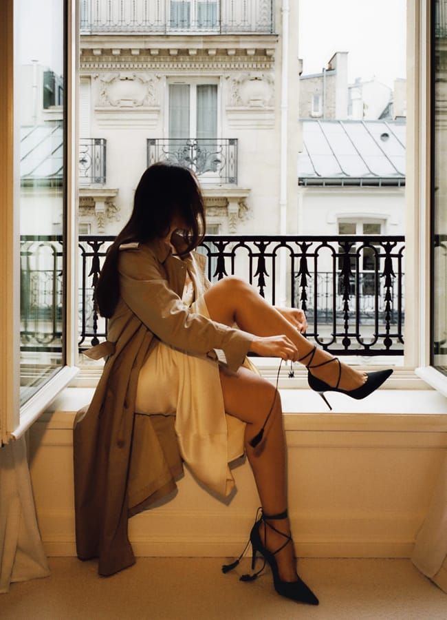 A woman in a trench coat sits on a windowsill, tying the strap of her sexy heels, with Parisian-style buildings visible outside the open window.