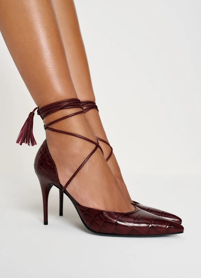 A close-up of a person wearing sexy heels—burgundy pointed-toe high heels with a quilted pattern and ankle wrap straps tied with tassels, against a plain white background.