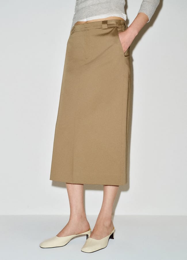 A person wearing a light grey top, a knee-length tan skirt with pockets, and white heeled mules stands against a plain white background, showcasing fresh new season fashion.