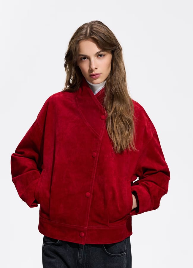 A woman with long brown hair wears a bright red suede jacket with buttons and pockets. This stylish piece is one of the season’s standout lightweight jackets. She stands against a plain white background with her hands in her jacket pockets, looking directly at the camera.
