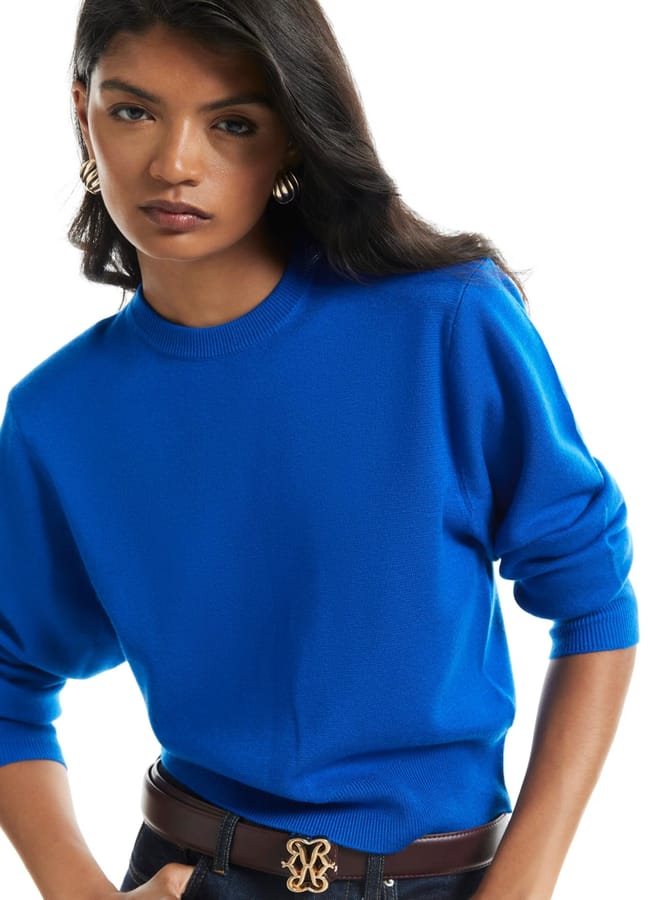 Woman with long dark hair wearing a bright blue jumper reminiscent of International Klein Blue, gold hoop earrings, and a brown belt with a gold buckle, standing against a white background. The bold colour of her jumper stands out everywhere she goes.
