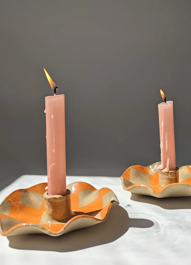Two pink candles in orange and cream chequered ceramic holders are lit, casting soft shadows on a white surface with a plain grey background—showcasing unexpected colour combinations that are making waves in current interior design trends.