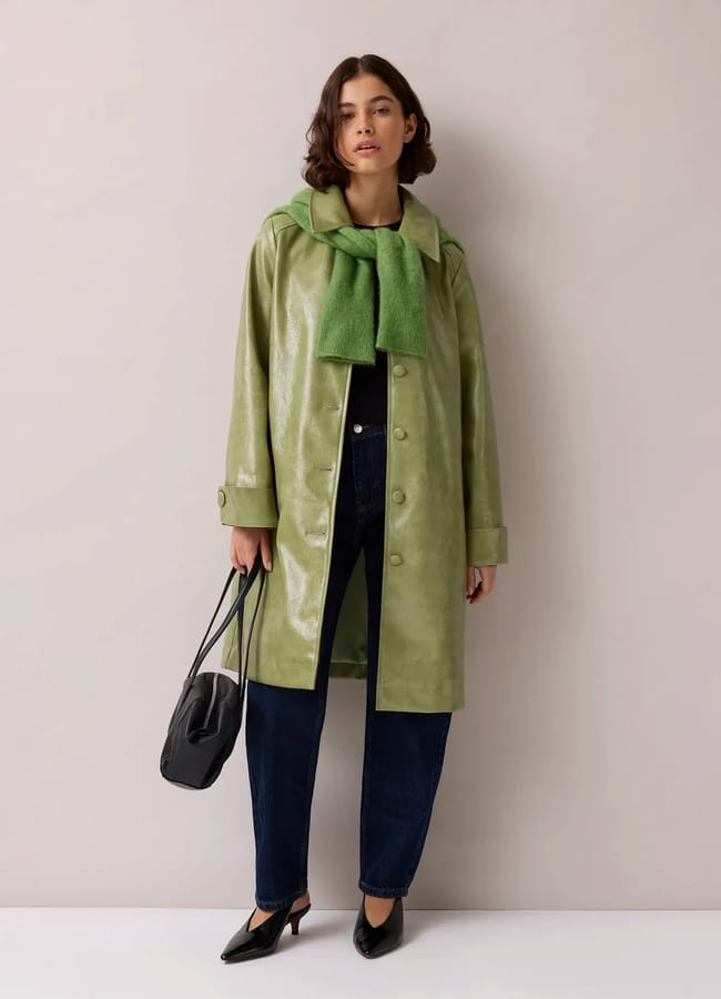 A woman stands against a light wall wearing a shiny green fashion coat, green jumper draped over her shoulders, dark jeans with a relaxed boyfriend fit, black heels, and holding a black handbag.