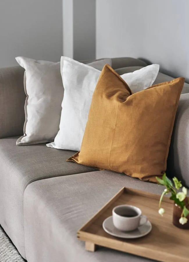 A beige sofa with two white cushions and one mustard yellow cushion, complemented by caramel homeware accents, sits next to a wooden tray holding a cup, saucer, and a small vase with flowers.