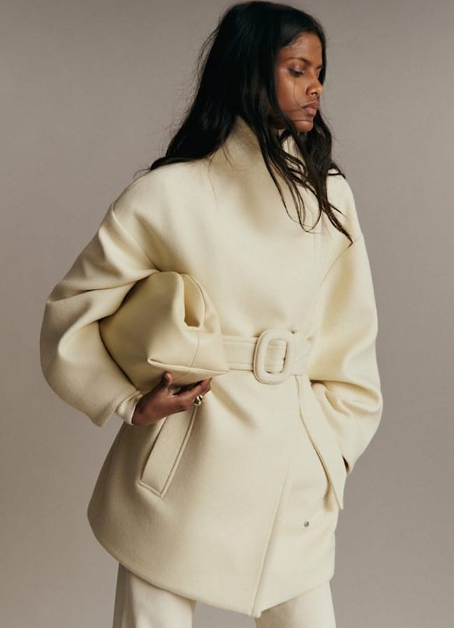 A woman wearing a cream-coloured belted coat—one of those timeless fashion essentials—holds a matching oversized clutch. She stands against a plain background, gazing to the side with a neutral expression.