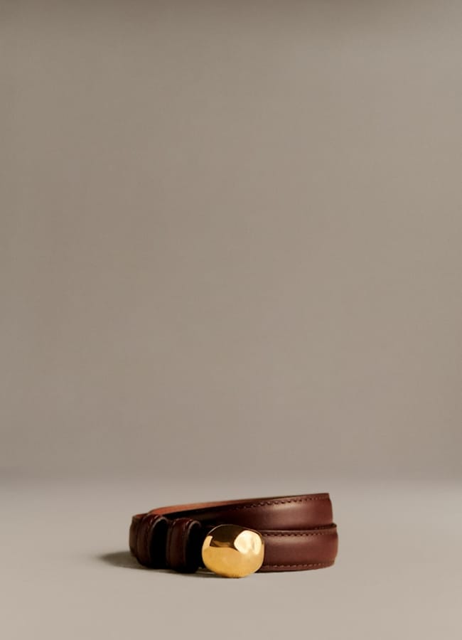 A brown leather belt with a gold oval buckle is coiled on a neutral beige surface, centred against a simple, plain background—an effortless wardrobe essential that can make everything look more expensive.