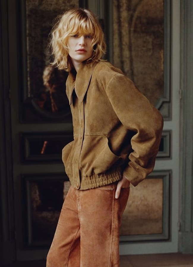 Person with wavy blond hair wears a brown suede lightweight jacket and rust-coloured trousers, standing with hands in pockets in front of a decorative door and mirrored wall.