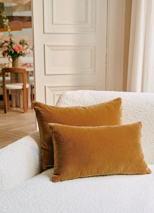 Two golden brown velvet cushions—perfect examples of caramel homeware—rest on a white textured sofa in a softly lit, elegant room. In the background, a wooden chair and a table with a floral arrangement are visible.