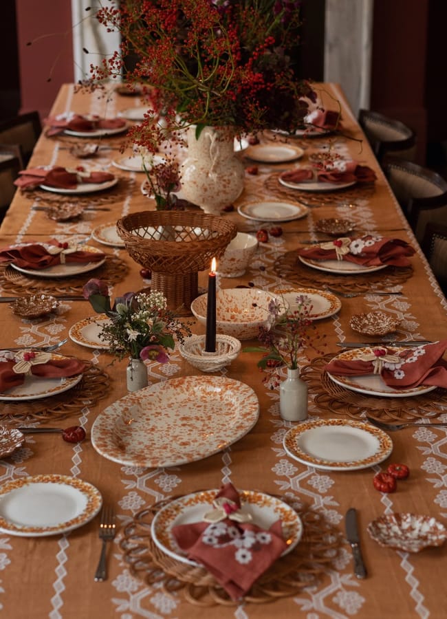 A long dining table set for a meal with vintage plates, woven placemats, rust-coloured napkins, a lace tablecloth, floral centrepieces, and a single lit candle. The table is decorated with delicate flowers, autumn accents, and touches of caramel homeware that add warmth and charm to the inviting setting.