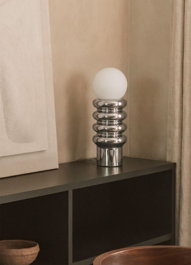 A modern table lamp with a round white bulb and a shiny, ribbed silver base sits on a dark cupboard next to a neutral-toned wall, abstract artwork, and a ceramic bowl—an elegant piece of chic homeware under £50.