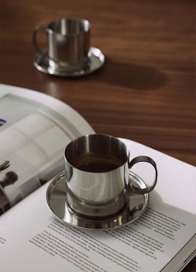 A silver cup with coffee sits on an open book beside a matching saucer—a perfect example of chic homeware under £50. Another similar cup and saucer are blurred in the background on a wooden table.