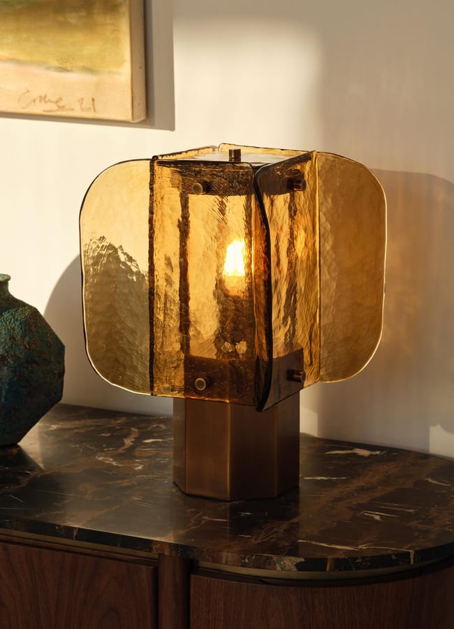 A modern amber glass table lamp with a square base sits on a dark marble-topped console table. Sunlight casts soft shadows on the wall. A textured green vase and a framed painting are partially visible nearby.