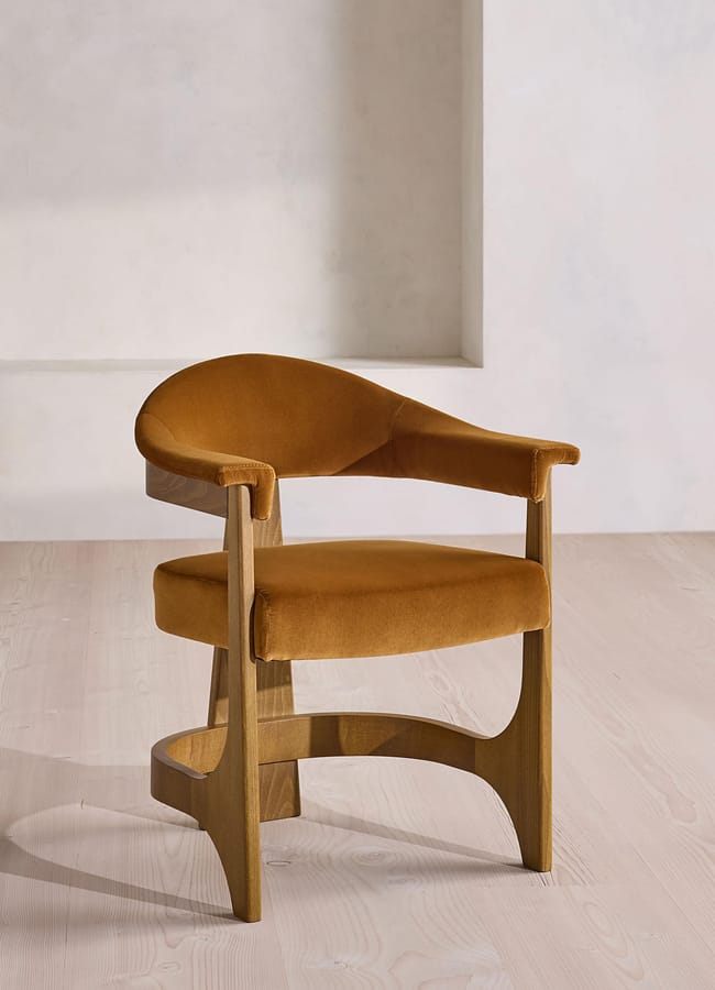 A modern wooden chair with a curved, mustard-coloured upholstered seat and backrest, reminiscent of contemporary caramel homeware styling, placed on a light wood floor against a light, minimalist wall.