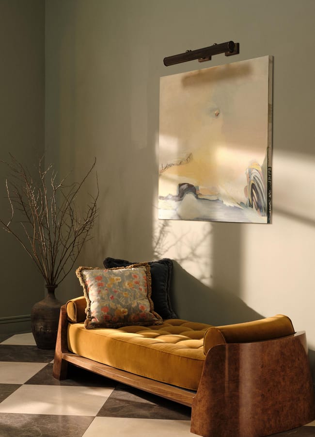 A mustard yellow daybed with two patterned cushions sits on a chequered floor. Behind it is a large vase with bare branches and an abstract painting on a light green wall, lit by soft sunlight.