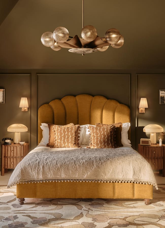 A cosy bedroom with a mustard velvet bed, decorative cushions, and touches of caramel homeware creates an inviting atmosphere. Two wooden bedside tables with lamps, wall sconces, framed art, and a modern chandelier add layers of style. The olive green walls and patterned rug complete the warm yet sophisticated look.
