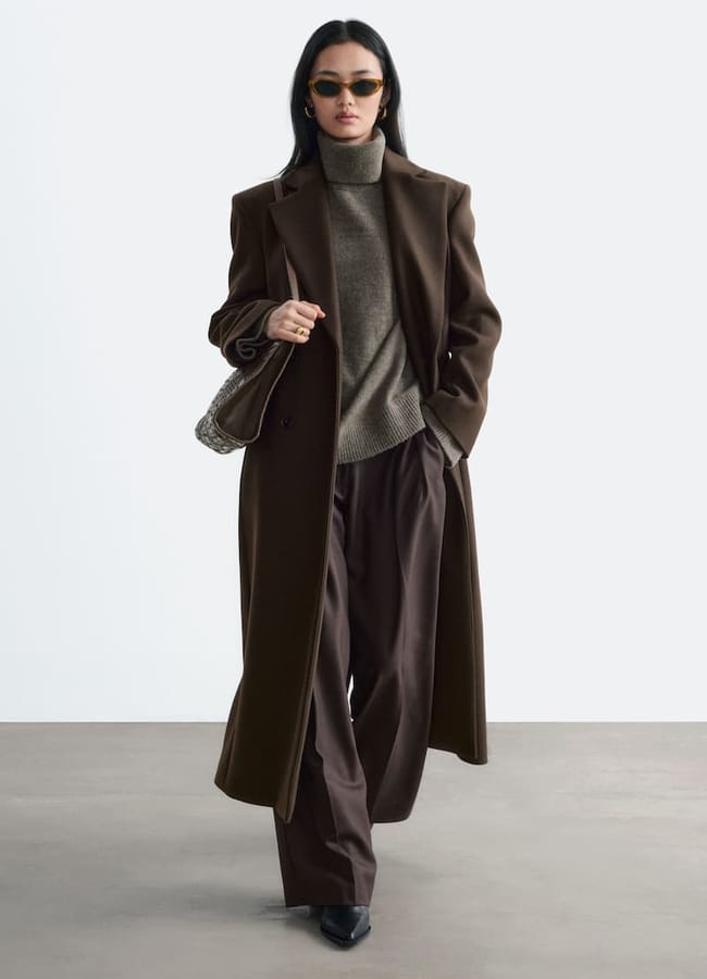 Woman in a brown overcoat, olive polo neck, and wide-leg trousers—a true showcase of wardrobe essentials—wearing sunglasses and heels, carrying a snake-print handbag, walking confidently against a plain background.