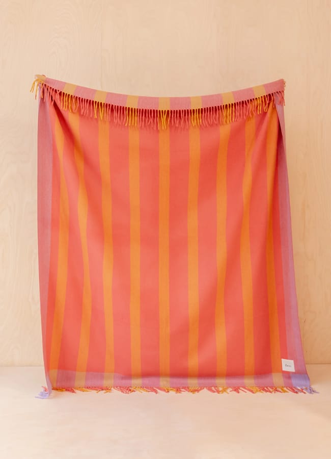 A woven textile with vertical orange and pink stripes—an example of unexpected colour combinations inspired by current colour trends—hangs against a light wood background. The fabric has fringe along the top edge and a small white tag on the bottom right corner.