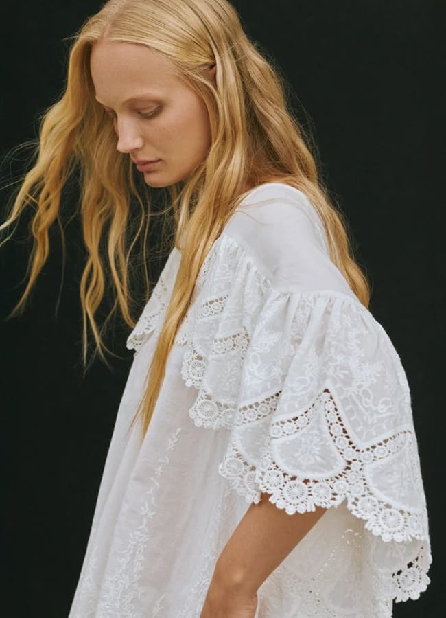 A woman with long blonde hair wearing a loose white dress from small fashion brands, featuring intricate lace and embroidery details, stands against a black background, looking down.