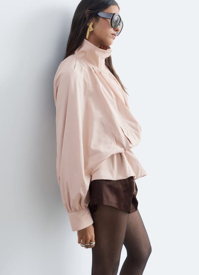 A woman wearing oversized sunglasses, a pale pink blouse with voluminous sleeves, brown shorts, black sheer tights, and one of this season’s must-have lightweight jackets stands sideways against a plain light background.