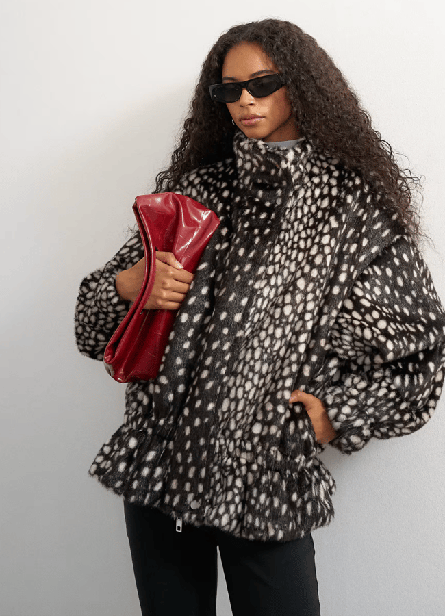 Woman with long curly hair wearing black sunglasses, a spotted black and white faux fur coat, and holding a large red clutch bag stands against a plain light wall—showcasing how cosy textures can be paired effortlessly with lightweight jackets for a chic layered look.