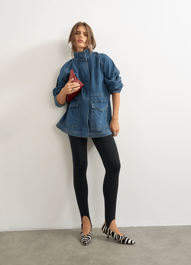A woman stands against a white wall wearing a blue lightweight denim jacket, black leggings, zebra-striped flats, and holding a small red clutch bag.