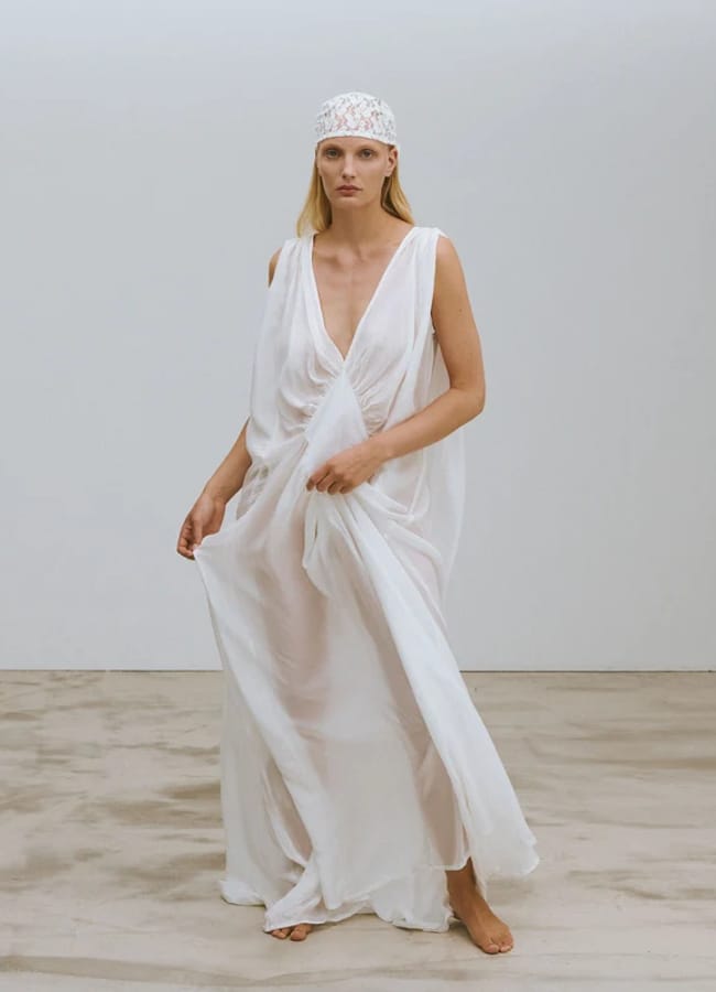 Woman stands barefoot on a plain floor, wearing a sheer, flowing white dress with a deep V-neck and a white lace head covering—an ethereal look that highlights the artistry often seen in small fashion brands—against a simple white background.
