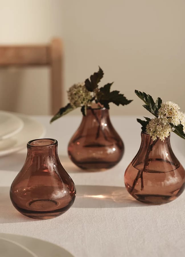 Three small, round, pinkish-brown glass vases—each a perfect example of chic homeware under £50—sit on a white tablecloth; two hold white flowers with green leaves, whilst the third is empty. A blurred wooden chair and plates are visible in the background.