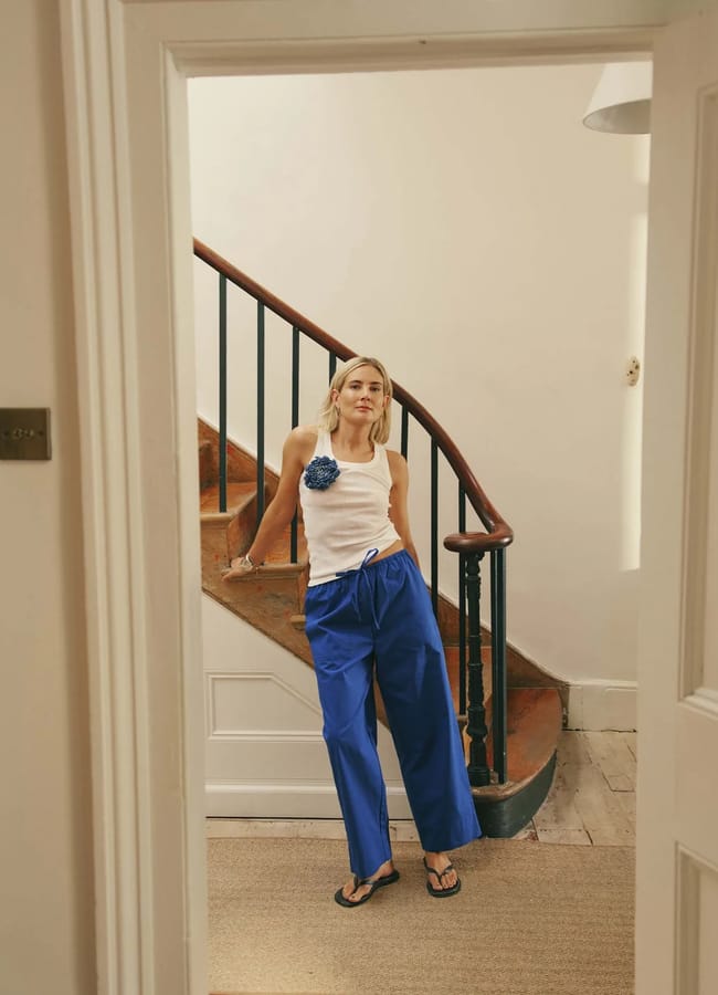 A woman stands indoors by a spiral staircase, wearing a white sleeveless top with a blue flower detail, bright blue trousers that echo the striking hue of International Klein Blue, and black flip-flops. She rests one arm on the railing and looks relaxed, as if colour is everywhere around her.