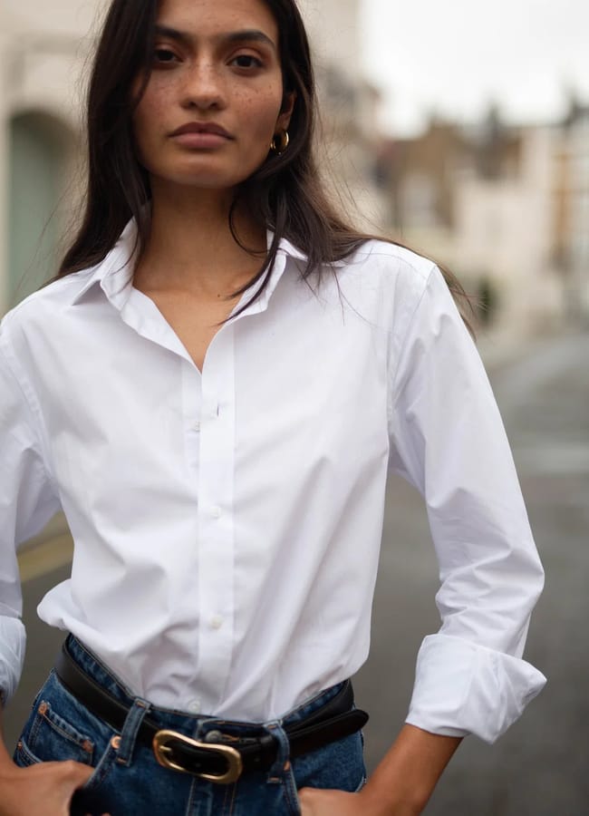 A person with long dark hair, wearing a white button-up shirt and blue jeans with a black belt—classic fashion essentials—stands outdoors on a city street with buildings blurred in the background.