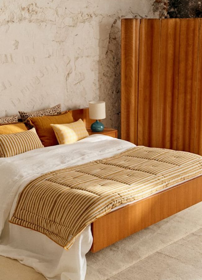A cosy bedroom with a wooden bed, striped bedding, mustard and white pillows, touches of caramel homeware, a bedside table with a turquoise lamp, and a wood room divider against a textured white wall.