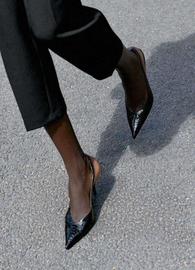 Person wearing sexy black pointed-toe slingback heels and black trousers walking on a textured grey pavement.