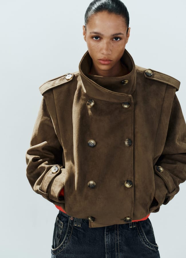 A person with slicked-back hair wears a brown double-breasted suede jacket—one of this season’s standout lightweight jackets with large buttons—and dark jeans, standing against a plain light background with hands in pockets.