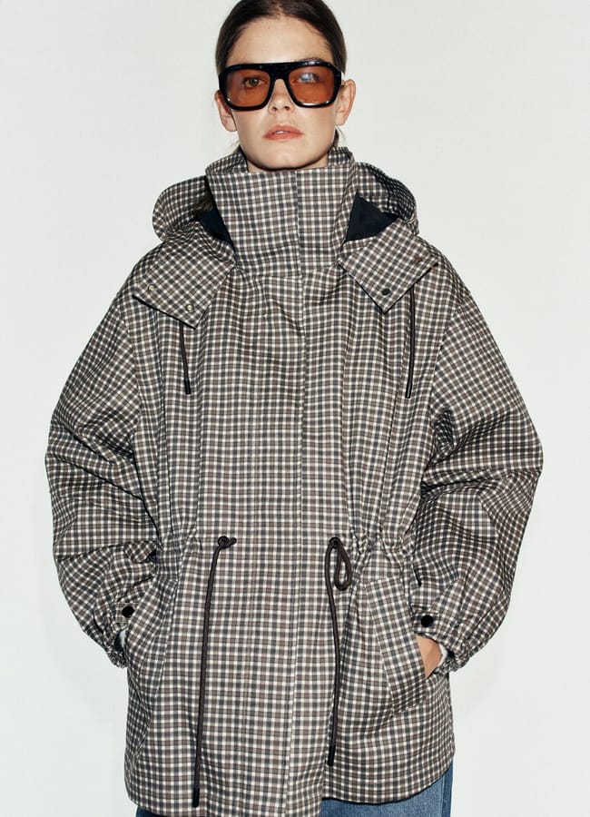 A person wearing oversized sunglasses and a brown and white chequered puffer jacket with drawstrings, perfect for those who love lightweight jackets, stands with hands in pockets against a plain white background.