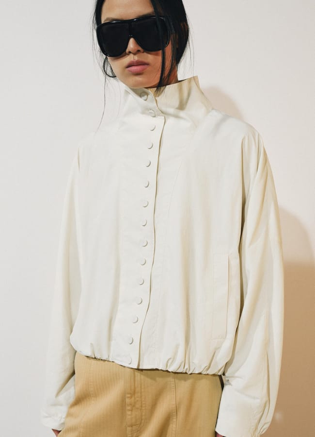A person wearing oversized black sunglasses, a high-collared white lightweight jacket buttoned up, and tan trousers stands against a plain light background.