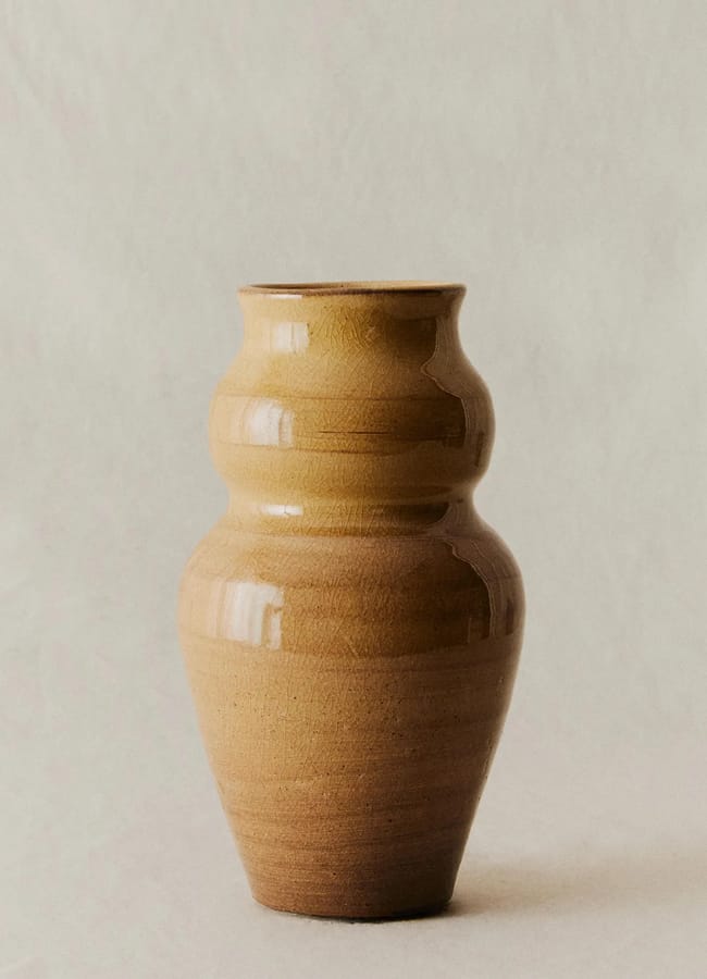 A brown, handmade ceramic vase with a rounded, tiered shape sits on a neutral surface against a light beige background, adding a touch of caramel homeware elegance to any space.