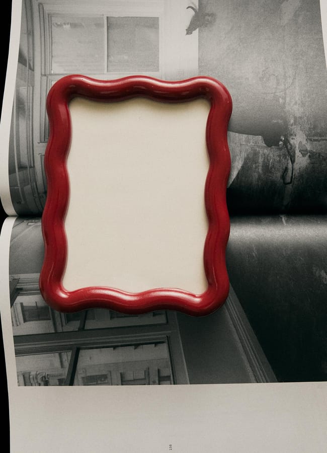 A red, wavy-edged empty picture frame—an example of chic homeware under £50—lies on top of an open magazine showing black-and-white photographs of rooms and a wall with a mounted animal head.